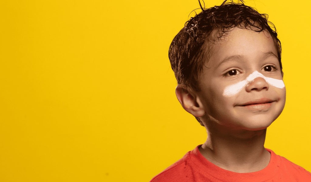 Menino, de aproximadamente 8 anos, com protetor solar aplicado nas bochechas e no nariz. Sua blusa é laranja e o fundo da foto é laranja.