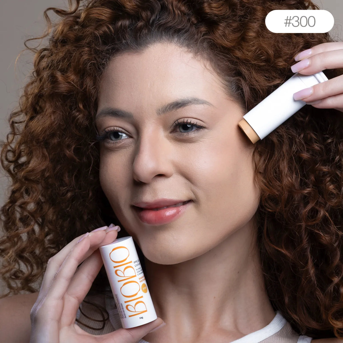Base Multifuncional em Bastão BioBio tom Castanha #300 sendo aplicada diretamente no rosto de modelo com pele média e cabelo cacheado, demonstrando a praticidade da aplicação.