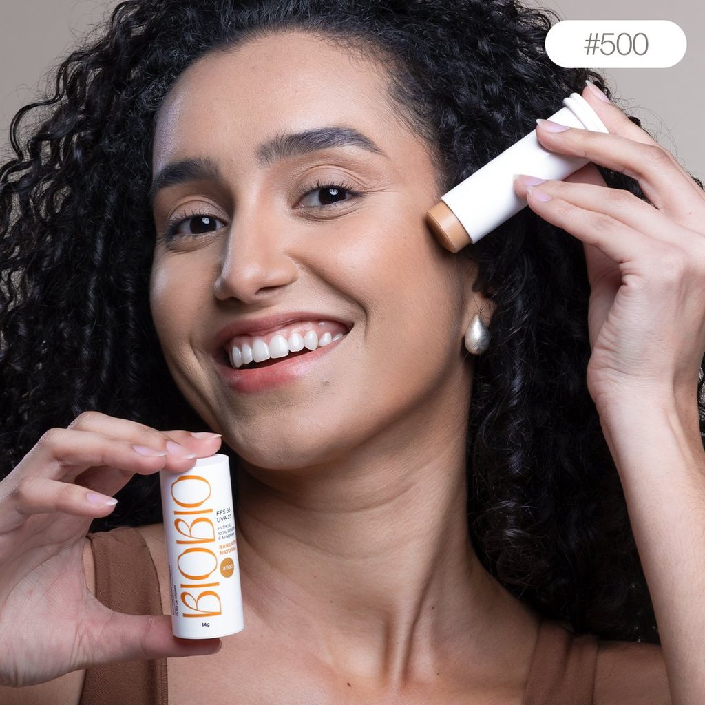 Base Multifuncional em Bastão BioBio tom Canela #500 sendo aplicada diretamente no rosto de modelo com pele média e cabelo cacheado, demonstrando a praticidade da aplicação.