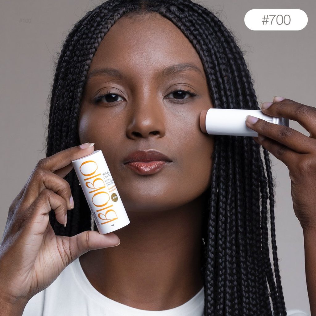 Base Multifuncional em Bastão BioBio tom Tâmara #700 sendo aplicada no rosto de modelo com pele negra e tranças longas, demonstrando a adaptação do produto a tons mais escuros .
