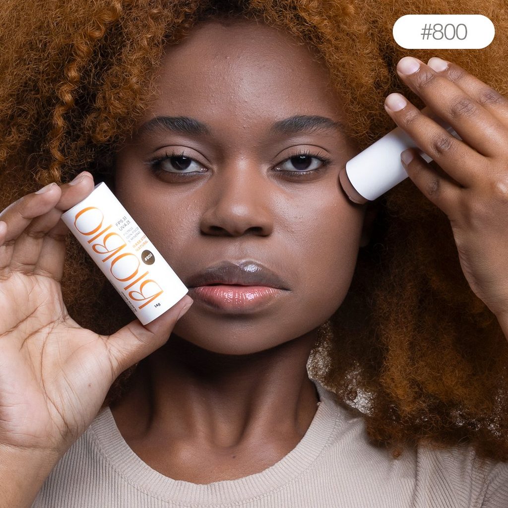 Base Multifuncional em Bastão BioBio tom Baru #800 sendo aplicada no rosto de modelo com pele negra e cabelo crespo, demonstrando a adaptação do produto aos tons mais profundos.