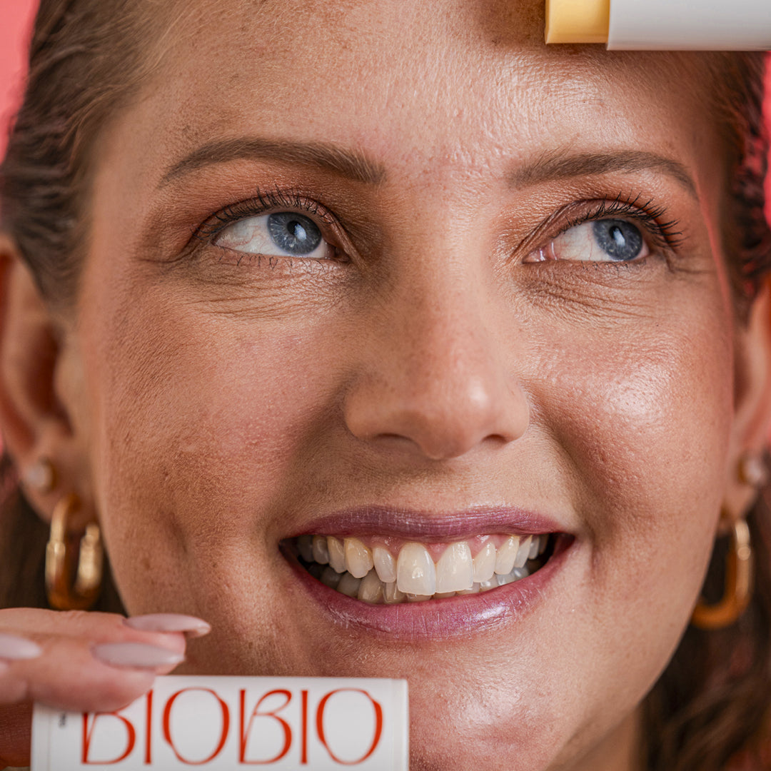 Close do rosto de uma mulher sorrindo e aplicando o Bastão Antimarcas BioBio na testa. Ela tem olhos claros e segura a embalagem do produto próximo ao queixo.