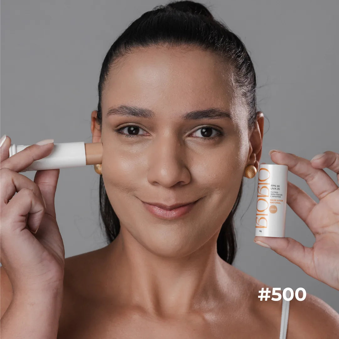 Base Multifuncional em Bastão FPS 33 da BioBio no tom #500 Canela: modelo aplica o bastão na bochecha e segura outro bastão de 14 g na outra mão.