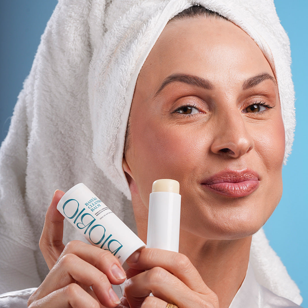 Mulher com toalha branca na cabeça segurando o Bastão Cleansing Balm da BioBio aberto. O demaquilante natural em formato stick facilita a limpeza facial, removendo resíduos de maquiagem e filtro solar. Ideal para rotina de double cleansing com produtos BioBio.