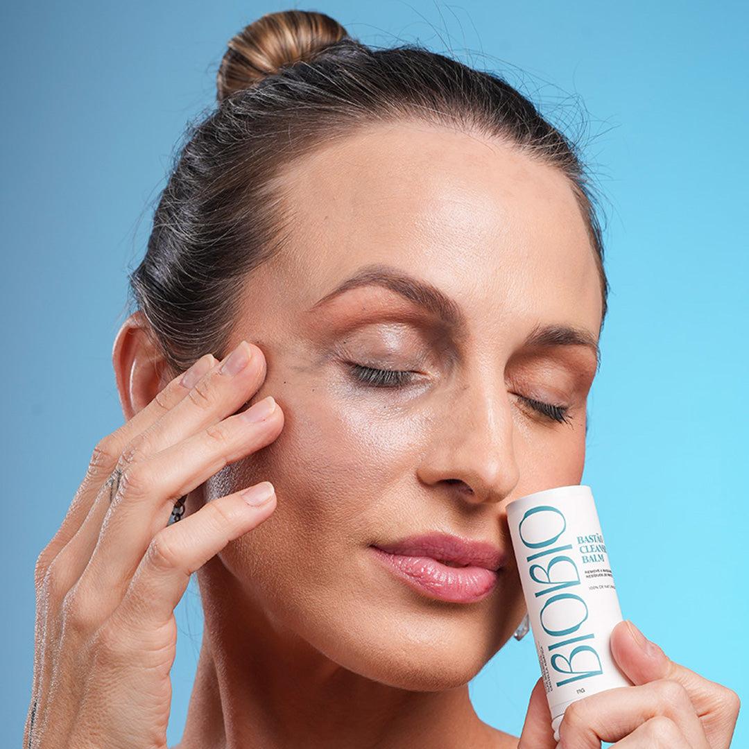 Modelo utilizando o Bastão Cleansing Balm da marca BioBio para remover maquiagem. A imagem destaca a praticidade do formato stick e a performance do demaquilante natural na rotina de skincare. Fundo azul claro reforça a identidade visual de frescor da BioBio.